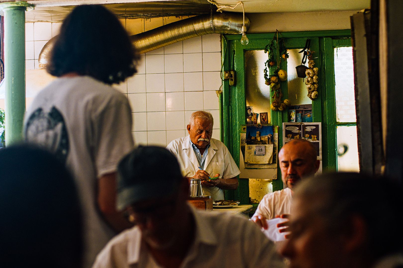 In a restaurant, the main subject at the center is an old man with grey hair and a moustache, the chef, taking note on a piece of paper. at the foreground we see blurry people eating and a man from the back standing. Behind him there is a green door with hanging garlic and a tiled white wall.