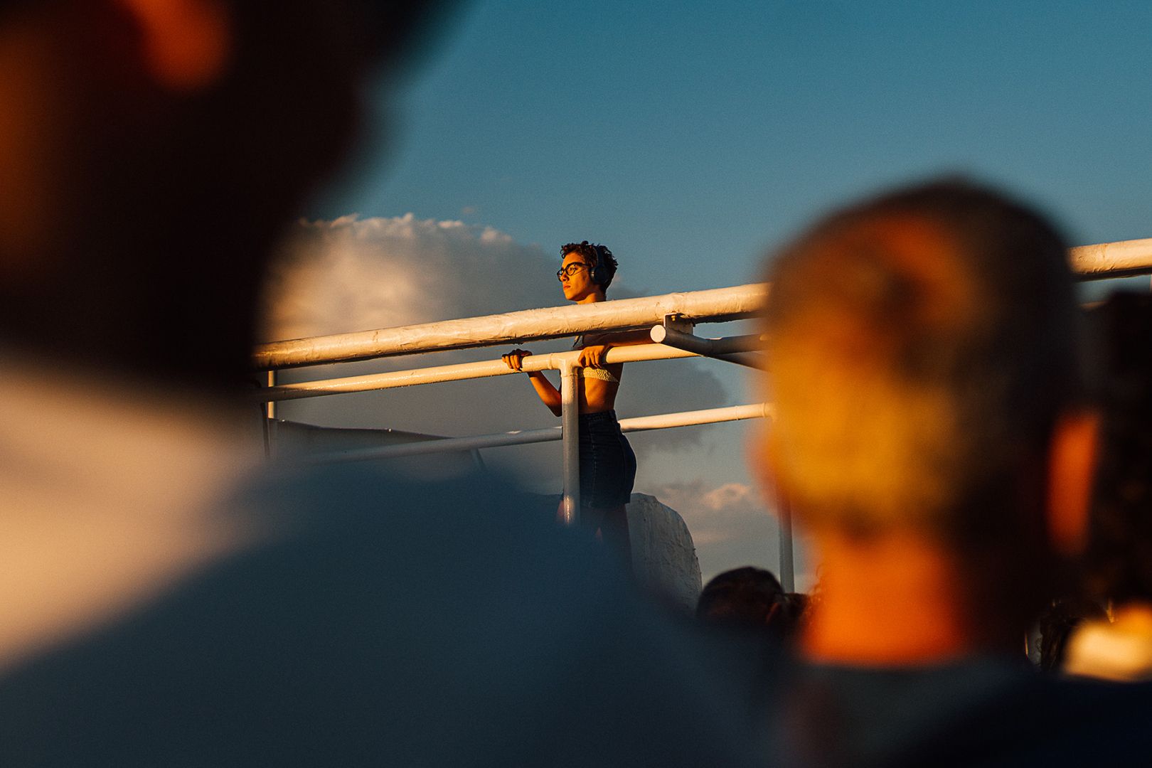 a person on a boat standing and looking at the horizon, lighted by the orangish sunset. At the foreground, 2 blurry heads of people from the back. We see the blue sky and a cloud in the background.