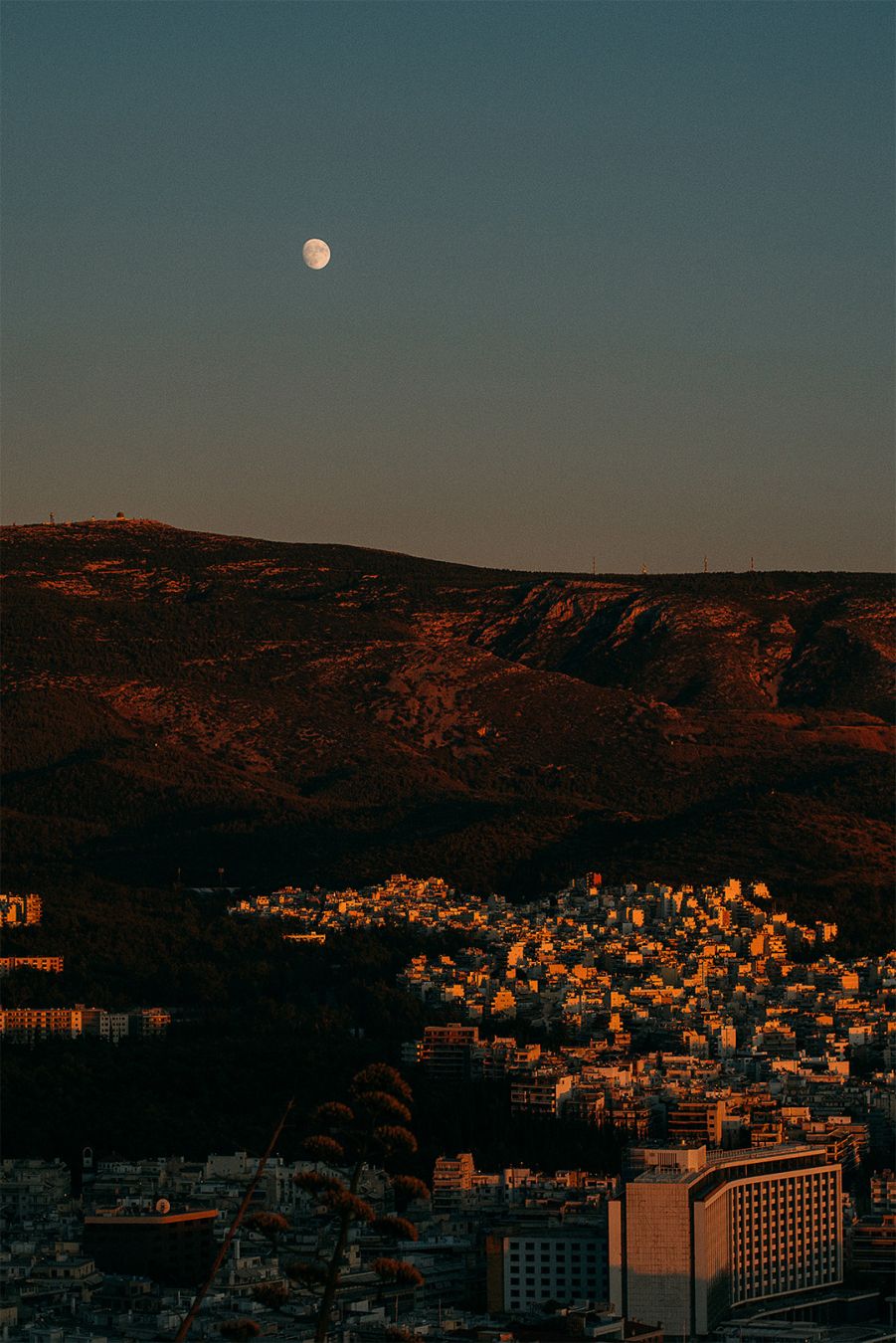 A landscape view of a city from a high point at the sunset. We see buildings illuminated by the sunset and a hill and the moon in the background.