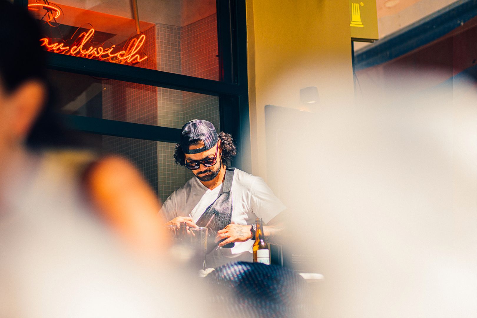 A man at a table of a restaurant wearing a white shirt, a cap and sunglasses, he is looking in his hip bag. In the top-left corner a led signage: "Sandwich". At the foreground: 2 highly blurred silhouettes.