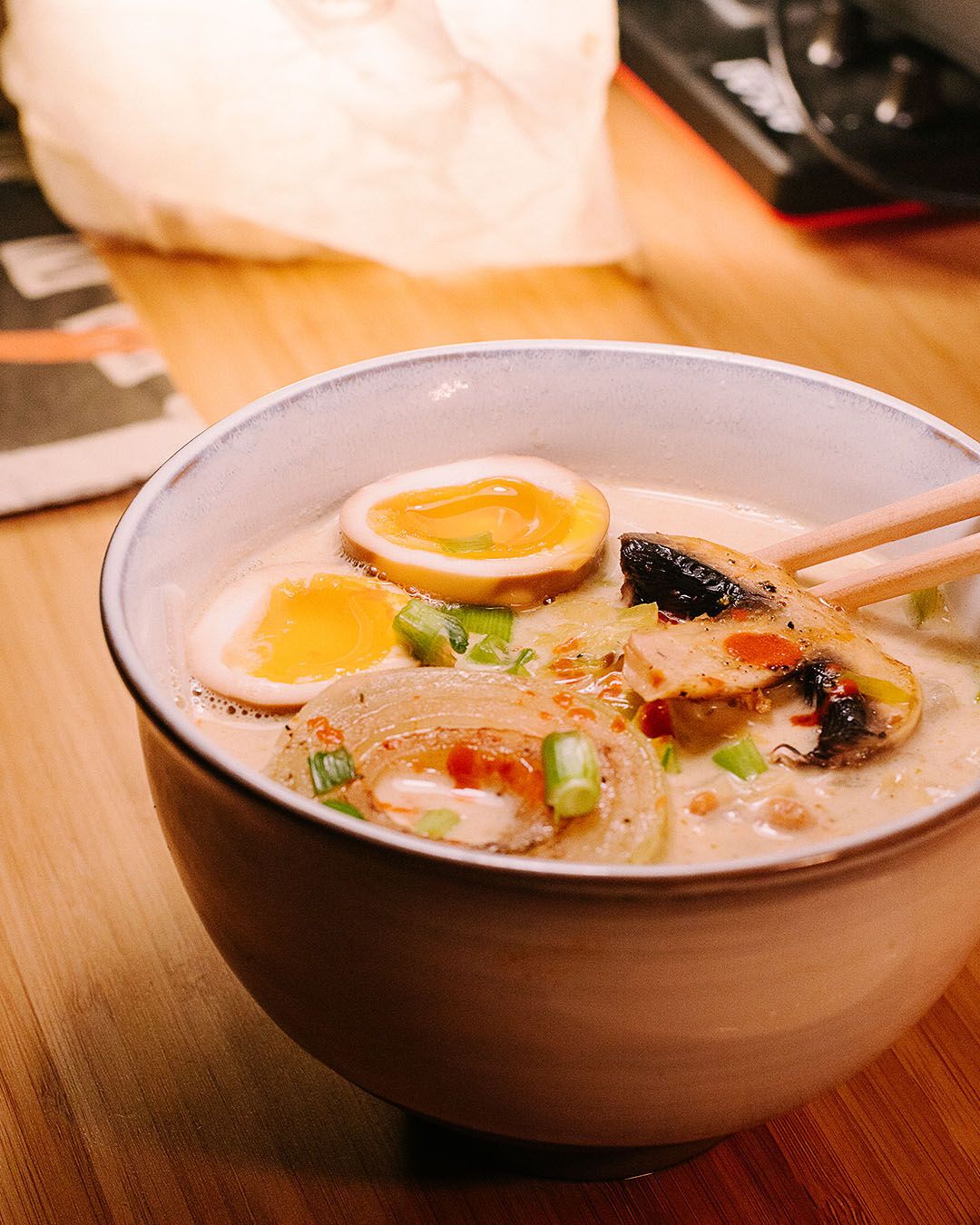 a close-up of an udon bowl on a wooden table. We see an egg cuted in half, a piece of mushroom, an onion slice, geen onions. The light is warm and cozy.