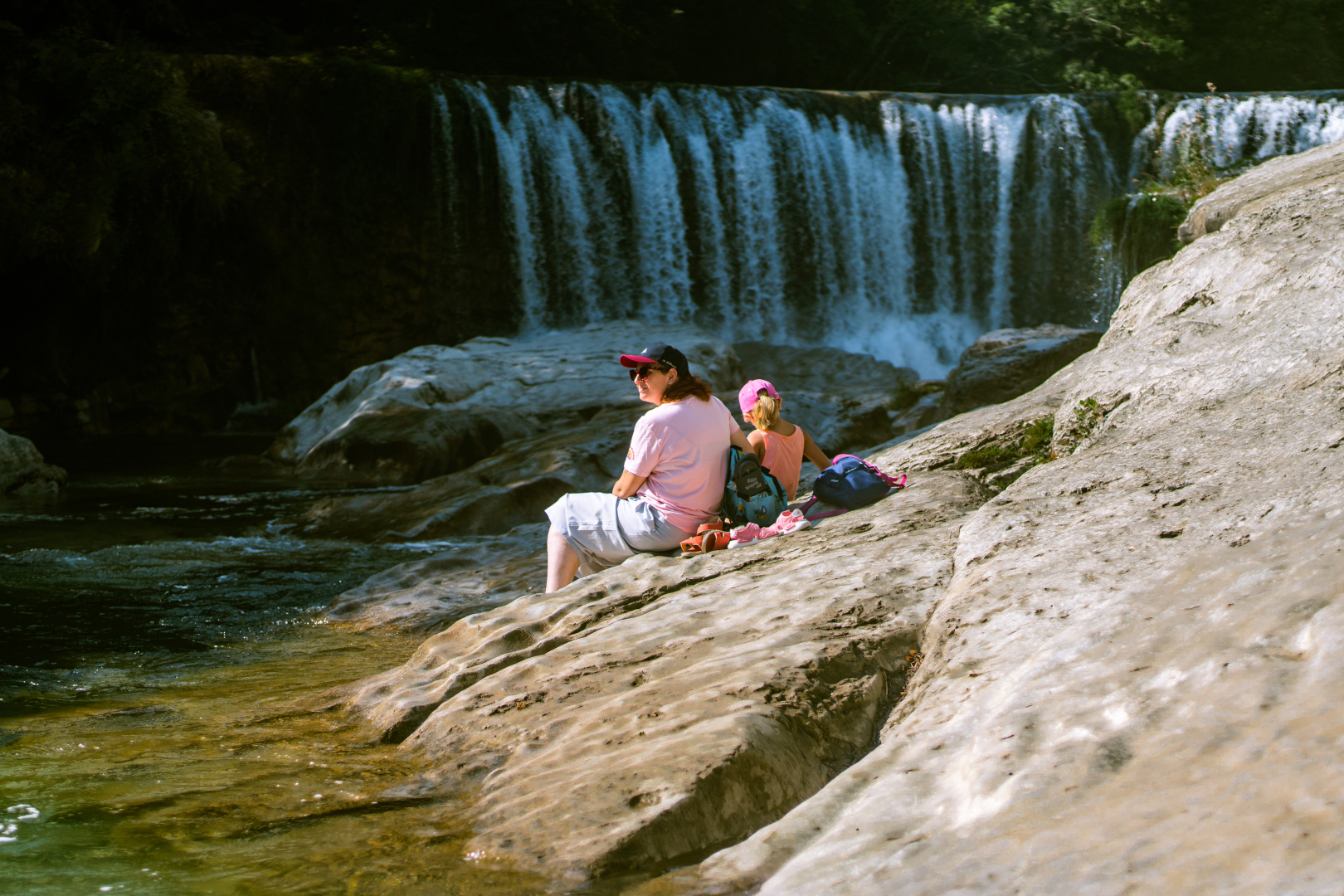 a woman and a girl seated on a rock near the water, from the back. A waterfall in the background. We see the face of the woman, lookink away from the waterfall, she is wearing a cap and sunglasses. Warm summer light.