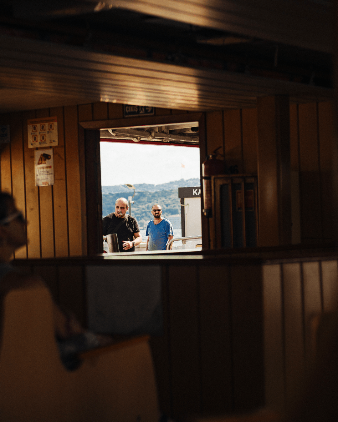 Photo taken inside a ferry boat, we see the passenger cabin at the foreground out of focus, some seats and a person sitted from behind. In focus, in the background we see two persons outside at the entrance the ferry (which is docked) One is smoking, the other one is looking toward the camera.