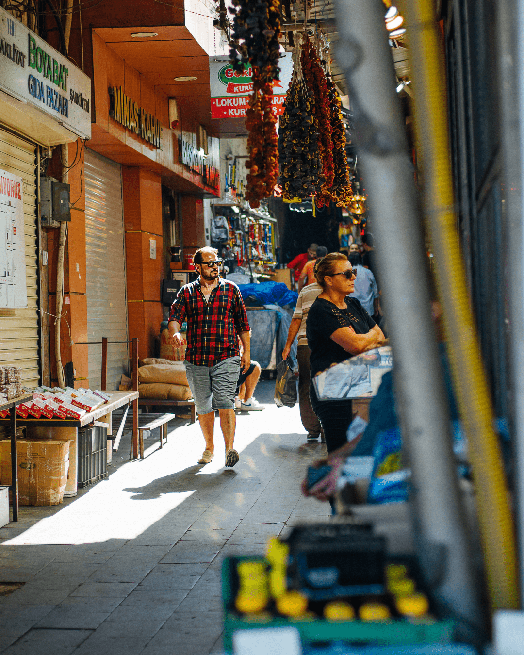 a narrow street filled with stores, we see a man walking towards the camera, he has a red plaid shirt and sunglasses. On the right side we see a woman arms crossed, waiting at a store front.