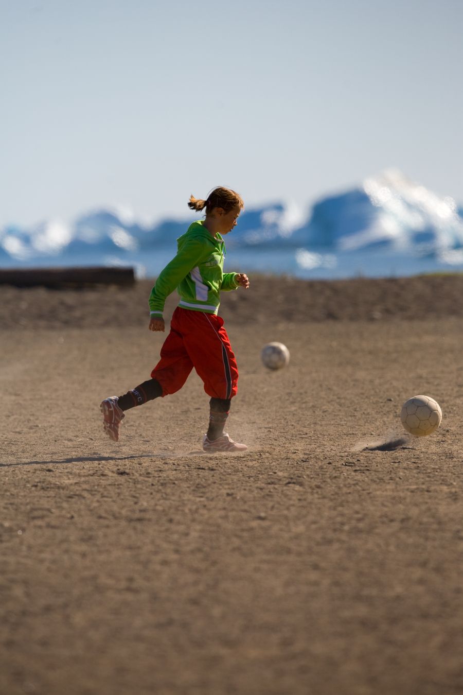 Une jeune fille, joue sur un terrain de foot sans herbe, elle soulève de la poussière dans sa course. Au loin on distingue des icebergs dans la baie de disko