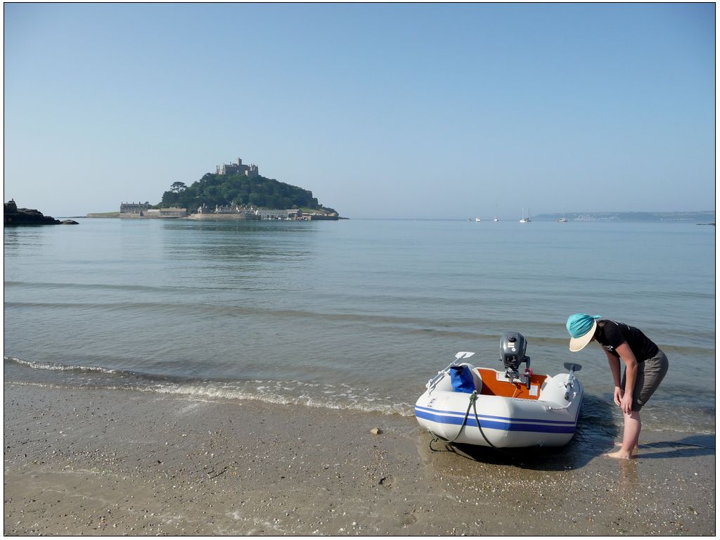 Photo de bord de mer. Au premier plan, le bord de l'eau où une annexe gonflable à moteur est échouée. Une jeune femme dont le visage est caché par un chapeau à large bord, remonte le bas de ses pantalons avant de pouvoir embarquer. 
À gauche de l'image, une île recouverte d'une forêt et surmontée d'un château est à quelques encablures du bord. La mer est toute lisse. On aperçoit au loin des voiliers au mouillage. L'ensemble de la photo est dans les tons bleus : Le ciel, la mer, l'annexe, le sac étanche dans celle-ci, le chapeau...