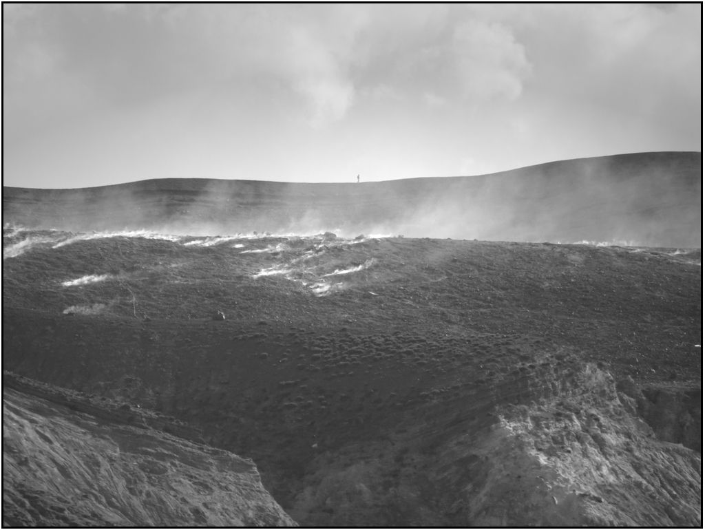 Photo Noir et Blanc de fumerolles sur une plaine volcanique. Au dernier plan et derrière la fumée, la silhouette minuscule d'un homme sur la crête se découpe dans le ciel.