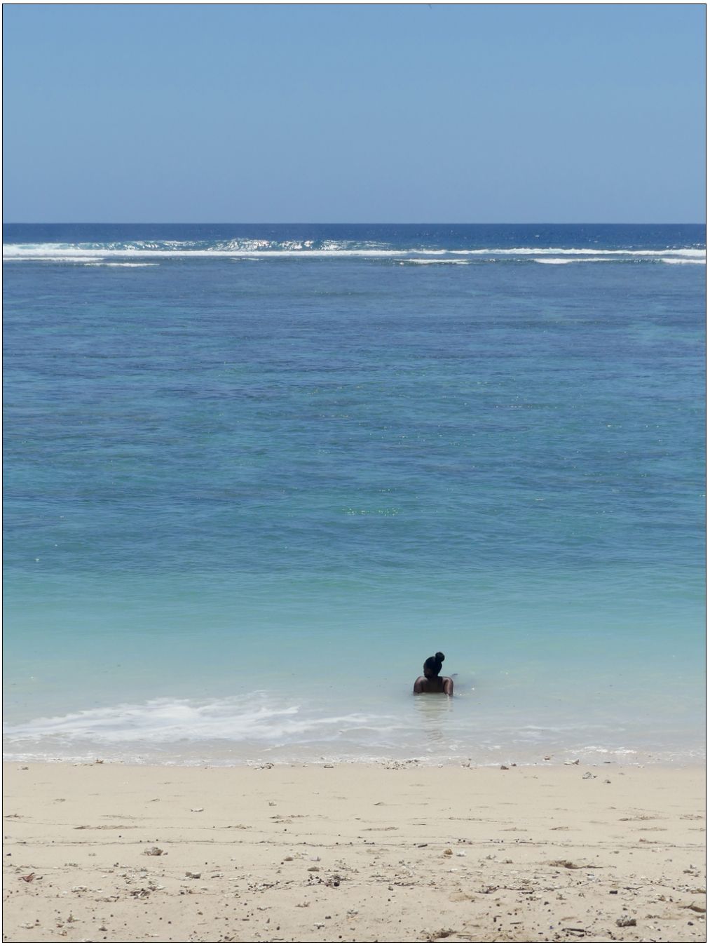 Photo de bord de mer. 
Au premier plan, la plage presque blanche forme une bande pâle. Le lagon forme un dégradé du bleu clair au bord de l'eau jusqu'à un bleu sombre à la limite du lagon où se forment les rouleaux. le ciel est lui d'un bleu uniforme.
Une seule silhouette allongée dans l'eau de dos. Seules les épaules et la tête sont visible. La coiffure en chignon se découpe sur la surface de l'eau.