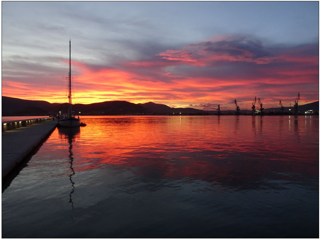 Photo d'un couché de soleil sur la mer. Le bas du ciel est orange, presque rouge. Cela se reflète sur la mer lisse comme un miroir. Sur la droite de la photo, un quai auquel est amaré un voilier. Au dernier plan, quelques grues et la montage se découpent dans le ciel.