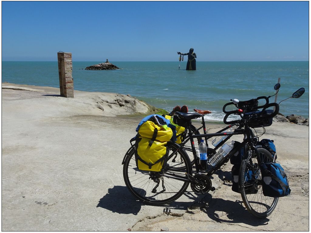 Payasage de bord de mer. Au premier plan, 2 vélos chargés de sacoches de voyages jaunes à l'arrière et bleues à l'avant. Au second plan, la mer dans laquelle se trouve une statue de déesse. Ciel bleu.