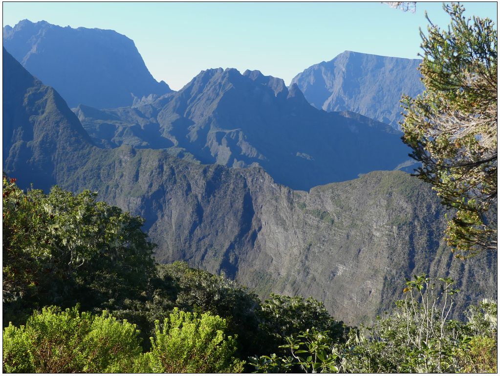 Photo du cirque de Mafate. Au premier plan, la végatation borde le bas de l'image et le côté droit. Des montagnes abruptes forment des falaises surmontées de crètes sur 3 autres plans.