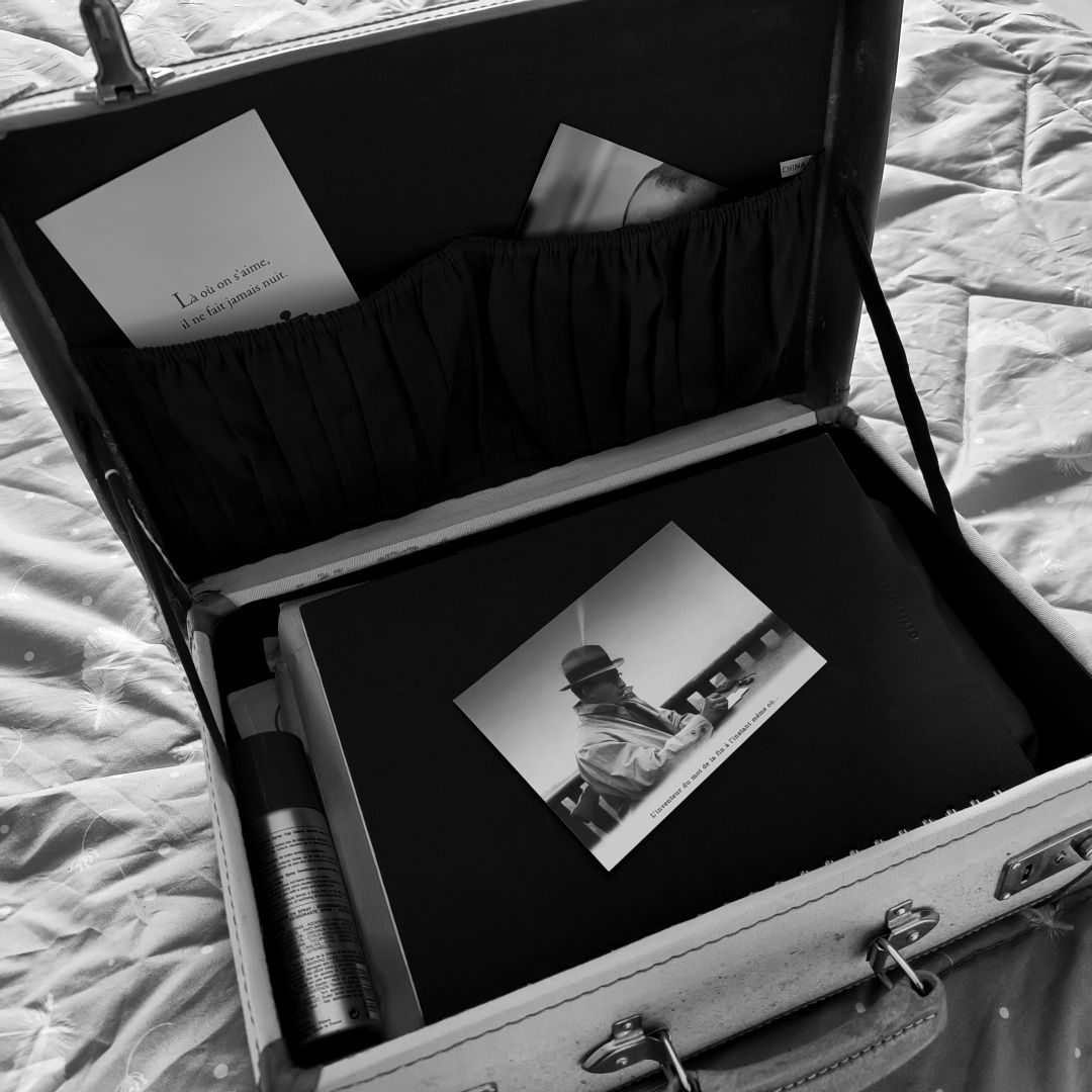 Photo en noir et blanc montrant une valise vintage, ouverte sur un lit. Dedans un spray, des documents noirs sur lesquels on voit une carte postale avec un homme qui écrit. Dans le couvercle, des poches dont dépasse une photo et une autre carte postale, à message.